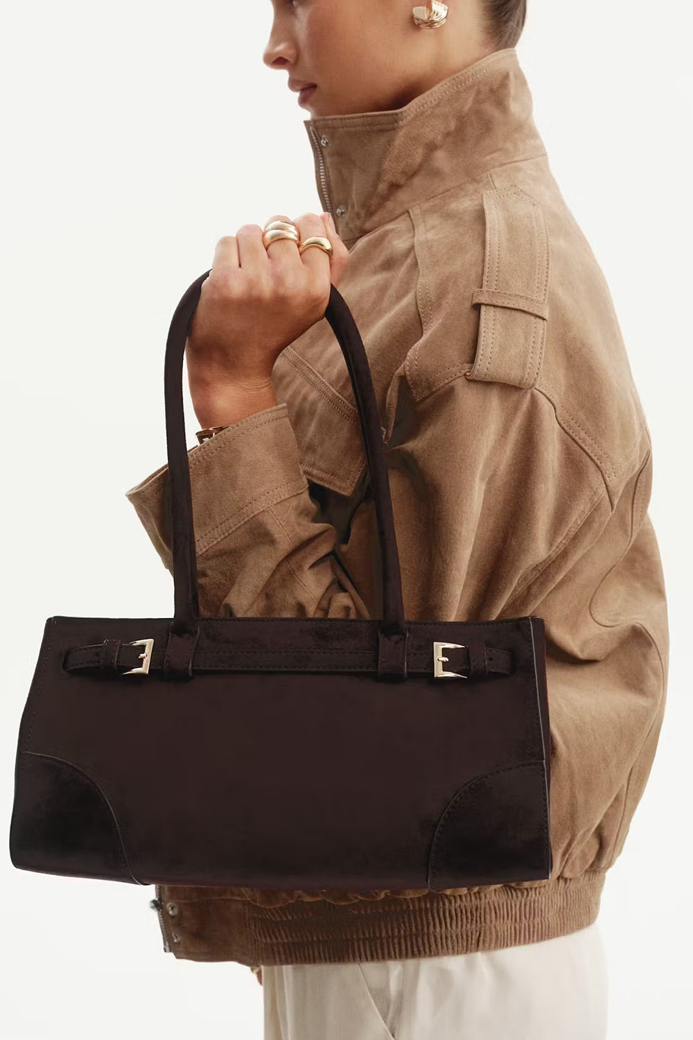 A person in a tan jacket holds a structured dark brown tote bag with double top handles and silver-tone buckle details against a clean white background.
